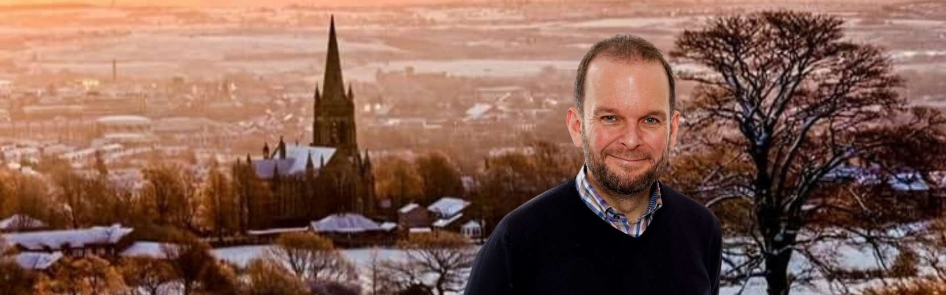  The image presents James Daly MP before a snow-draped Walshaw backdrop. His attire is casual with a dark sweater over a checkered shirt. The winter scene behind him showcases a church spire amid the snow-blanketed village, under a warm-hued sky.