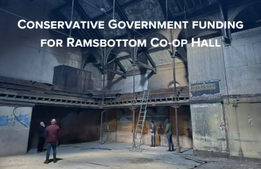  The image shows the Ramsbottom Co-op Hall's interior, characterized by vintage architectural details and a balcony, in need of restoration. Visible signs of wear, such as peeling paint, indicate neglect. People in the hall suggest a planning or assessment activity. The text highlights Conservative Government funding for the hall's renewal. Generated automatically.